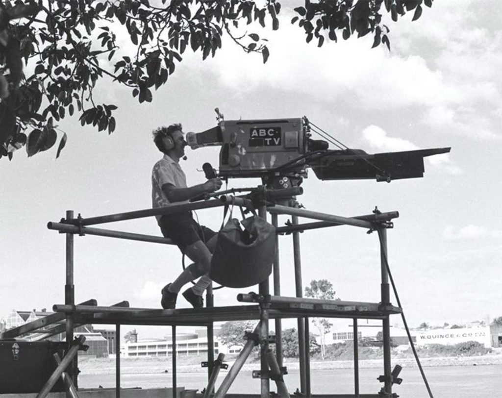 Black and white photo of ABC camera operator seated at large broadcast camera and tripod, on scaffolding beside Brisbane river
